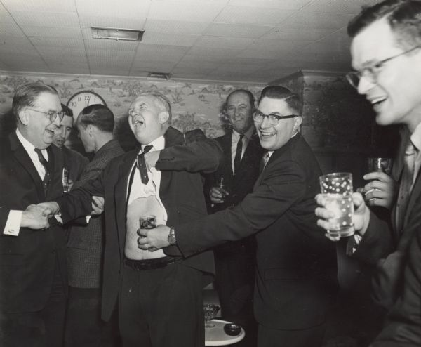 Sid (center) is hoisting up his shirt and exposing his stomach in a roomful of male colleagues. One man standing near him is holding a drinking glass with ice against Sid's stomach as other colleagues look on with drinks in their hands.