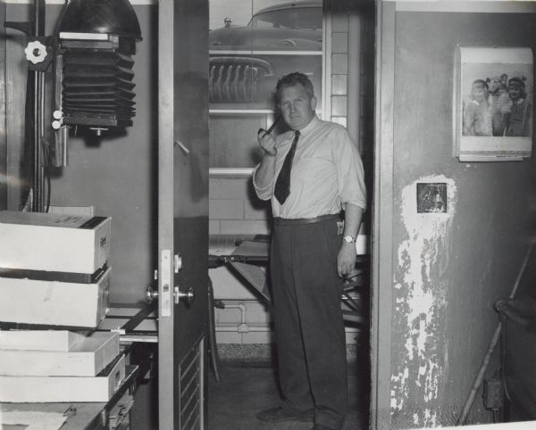 Sid standing in a print drying room adjacent to a darkroom (likely at Gisholt). He wears trousers and a tie, and is holding his pipe in his right hand. A parked car can be seen through a window in the background.