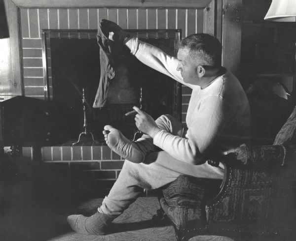 Sid, wearing thermal underwear, is sitting in an upholstered chair in front of a fireplace, and holding his sock aloft. He is holding a cigar in his left hand.