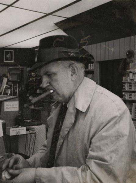 Sid, wearing an overcoat, hat, and with a cigar in his mouth, standing in a photography store.