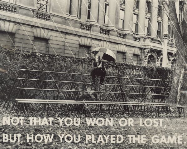 Sid, holding an umbrella and sitting on bleachers outside the Wisconsin Historical Society building. Rain (graphite lines) is annotated on the negative. At the bottom is the text: "Not That You Won Or Lost, But How You Played The Game."