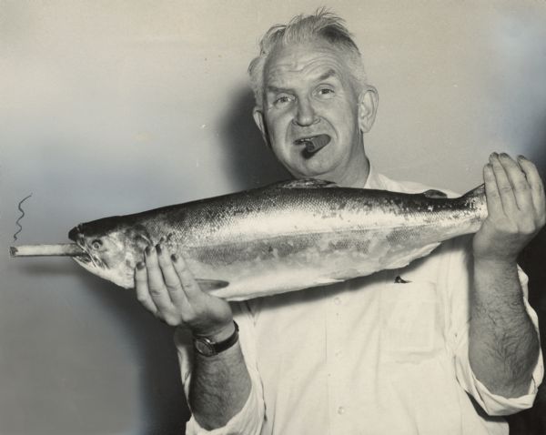 Waist-up portrait of Sid with an unlit cigar in his mouth, holding up a trout in both hands. The trout has a cigar stuck in its mouth. The area around the trout's mouth, as well as the tail area, has been airbrushed. A trail of smoke has been added to the cigar in the trout's mouth. Sid's nails are likely stained with pyrogallol, a staining agent used in the development of film. 