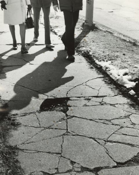View of the 2800 block of Atwood Avenue showing the condition of the sidewalk. Show three sets of people's legs walking toward a large hole and cracks in the sidewalk.