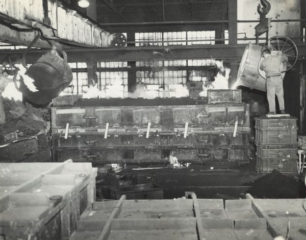 Interior view of Gisholt Machine Company. A man is standing on metal parts and pouring, what is probably molten metal, from a vat into a mold or industrial container. Another man is standing in the background with his hands on his hips in front of the large factory windows.