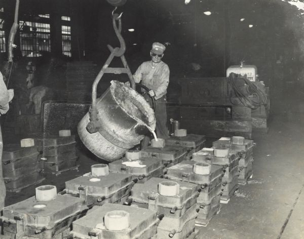 A Gisholt Machine Company factory worker, wearing protective glasses, is using a wheel to position a large cauldron for pouring molten metal into a mold to cast parts. Other workers are in the background.