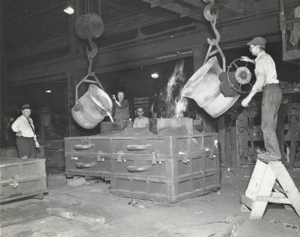 Two employees of Gisholt Machine Company are pouring molten metal from large cauldrons into molds as another worker looks on. A company supervisor is standing on the left.