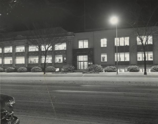 View at night of 1245 East Washington Avenue towards the main entrance of Gisholt Machine Company. The sign above the entrance reads: "Gisholt Machine Co." Lights are on inside the factory offices. There is a car parked along the curb in the foreground, and snow is on the ground. The company spanned the blocks of East Washington from 1245 to 1301. 