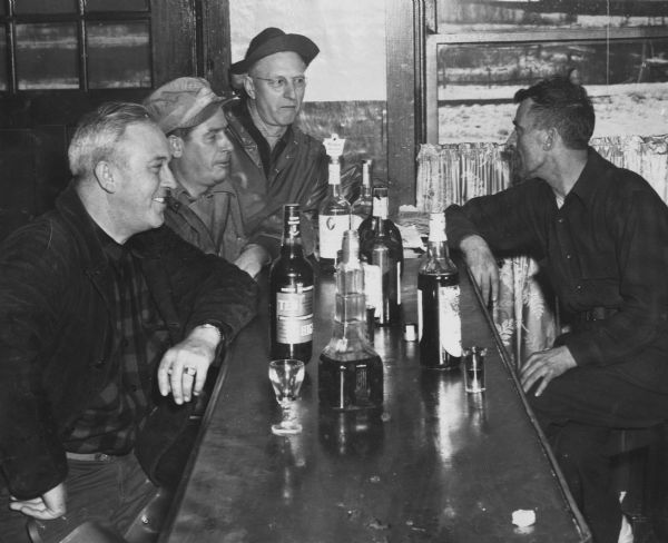 Three men sit at a bar socializing with a bartender and drinking shots. The man in the foreground is holding a cigarette. Various bottles of alcohol are in front of them. 