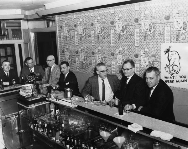 View of Gisholt employees, all men, wearing suits and neckties, sitting around the bar of an unidentified tavern. On the wall on the right is a Sid Boyum cartoon, which depicts a bird with its head in the sand with the caption: "What! You here again?" 
