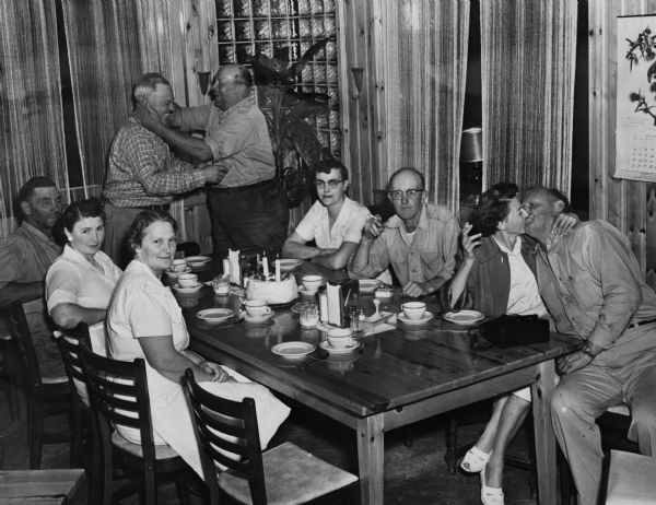 Group portrait of men and women sitting at a table in the corner of a room, perhaps a restaurant, at a birthday party. There is a cake with candles in the middle of the table. In the background two men are standing and hugging, and on the far right a man and a woman are kissing. Three of the woman are wearing white dresses, which may be uniforms.