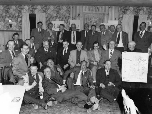 Group portrait of the Ice Chippers, members of a winter ice fishing group based in Madison. They are posing indoors, and Sid Boyum is sitting on the floor in the center holding a cigar. On the right, a man is holding up an Ice Chippers poster.