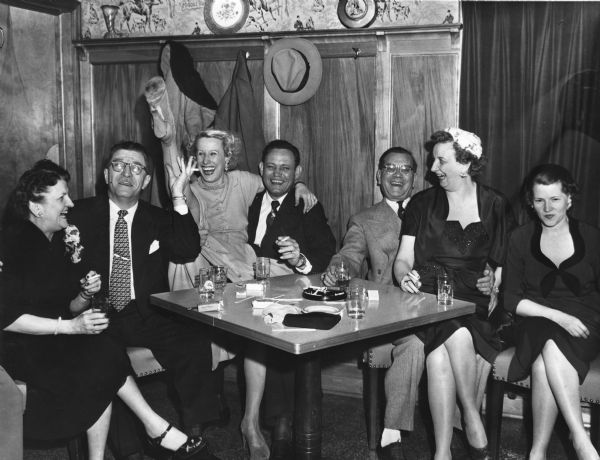 A group of men and women sitting around a table smoking cigarettes and gesturing, probably at a bar or restaurant. One of the women is kicking her leg high up in the air. There are drinks and an ashtray on the table in front of them.