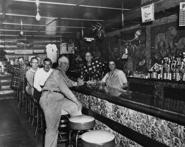 Group portrait of men, including Sid's friend, Eugene Beckman (in front), sitting on stools and drinking at a fishing-themed bar. A man and a woman are standing behind the bar.