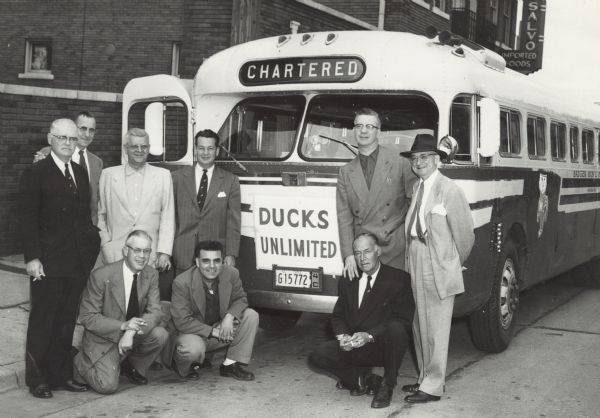 Group portrait of men in front of a chartered Badger Bus Lines bus, with a Ducks Unlimited sign taped on the front. The bus is parked in front of a brick building, and a sign in front says, in part: ". . . SALVO Imported Goods."