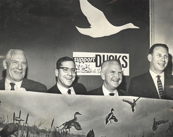 Group portrait of four men, taken at the Club Chanticleer, holding up a banner depicting ducks in a marsh. There is a "Support Ducks Unlimited" poster in the background underneath a silhouette of a duck flying. L to R: Ken Harley, charter member; Joseph Steuer, Sid Boyum, Herbert Welsh.