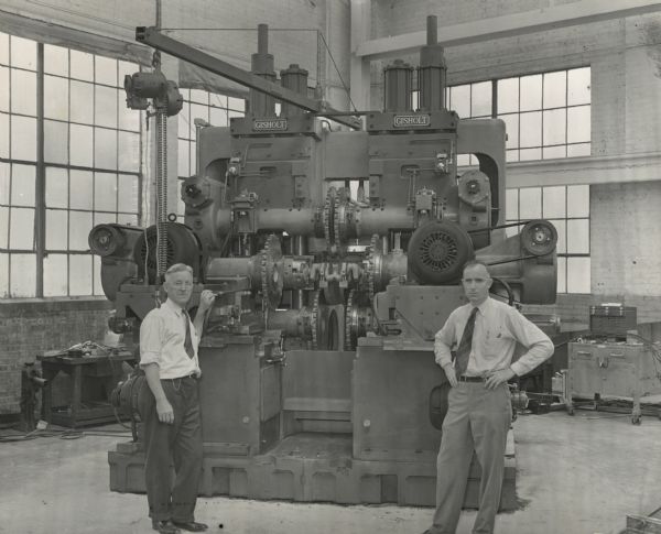 Two men in button-down shirts and neckties are standing in front of a large metal tooling machine in a corner of the Gisholt Machine Company factory. Large windows are in the background.