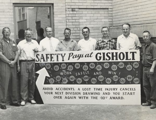 Workers posing outside a factory building at Gisholt Machine Company by a large sign for an employee contest that reads: "Safety Pays at Gisholt." The sign promotes safety with cash rewards. The workers are: (left to right) John Weston, Sid Boyum, Roy Langdon, Floyd Pavey, Steve Connor, James Yahn, Elmer Pederson, and Earl Turner.