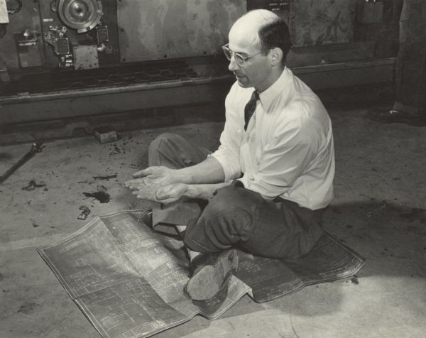 A male office worker wearing a white shirt and necktie is sitting cross-legged on a blueprint drawing on the factory floor at Gisholt Machine Company. The man is holding his palms open in front of him. There is a large piece of machinery in the background.