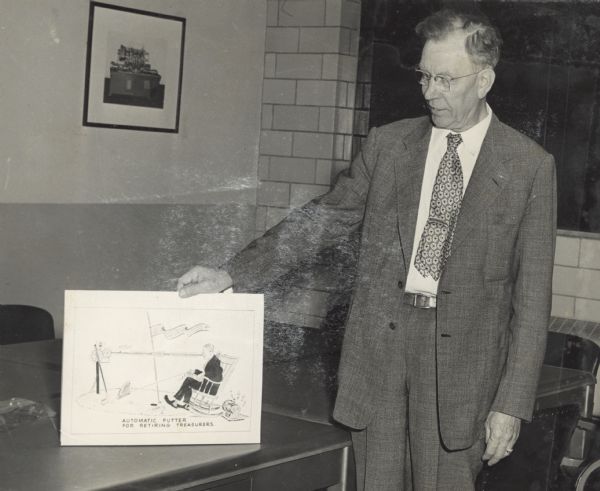 The Gisholt Machine Company treasurer is standing and displaying a Sid Boyum drawing on top of a desk. The drawing was created for the treasurer in celebration of his retirement, and depicts the treasurer sitting in a rocking chair on a putting green using an automatic putter.