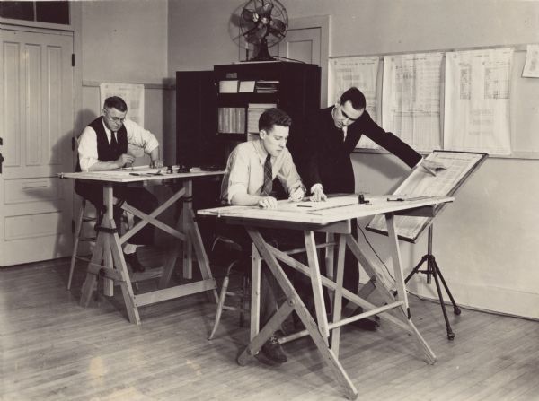 Two draftsmen in shirtsleeves working at drafting tables. A man is standing near the man in the center. What appear to be charts and a bookcase are in the background in a corner of the room.