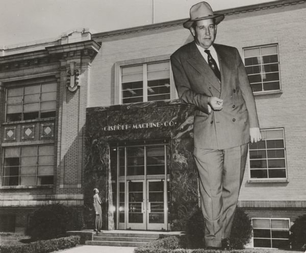 Composite image of a man wearing a suit standing in front of the Gisholt Machine Company entrance. He is looking up at a man scaled as tall as the building, and who is leaning against the front doorway.