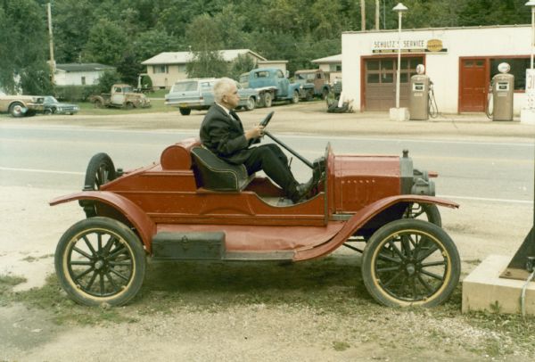 Sid Boyum sitting in a parked, red Model-T outside Moody's Museum in McGregor, Iowa. Sid is wearing a suit and bow tie, and has a cigar in his mouth. Across the street behind him is Schultz's Service station, with parked automobiles and trucks in its yard. Tree-covered bluffs and houses are in the background.