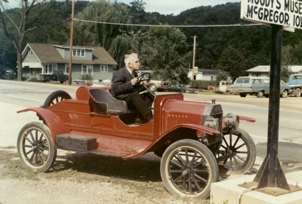 Sid Boyum sitting in a parked, red Model-T outside Moody's Museum in McGregor, Iowa. He is wearing a suit and bow tie, and has a cigar in his mouth. Tree-covered bluffs are in the background.