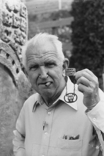 Waist-up portrait of Sid Boyum in his backyard proudly holding a medal he won for an entry to the Burlington Liars Club in 1976. The entry read: "During a recent cold snap, I saw a nightcrawler steal the fur coat off a caterpillar and crawl back in his hole." The medal is a visual pun for the club, fashioned into the shape of a lyre, which is centered in a ring that suspends from two short chains attached to a miniature plaque that reads "World Champion Liar."

Sid holds a cigar in his mouth, and wears a collared shirt with breast pocket full of cigars and a pair of eyeglasses. A small microphone is attached to a shirt button for what must have been an interview over his award.

