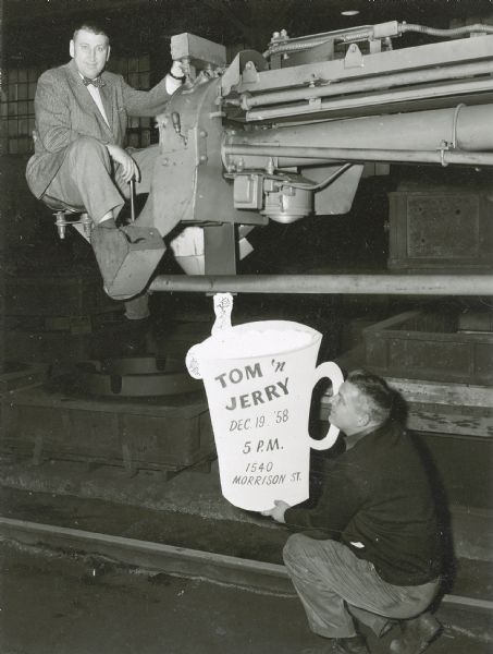 Two male employees of Gisholt Machine Company advertise an upcoming holiday party at 1540 Morrison Street. One of the them, formally dressed in suit and tie, is in the operator seat of a heavy piece of machinery. The other squats in the foreground, holding a sign in the shape of a mug with the time, date and address for the "Tom 'n Jerry" party, named for the popular cocktail at Christmastime.