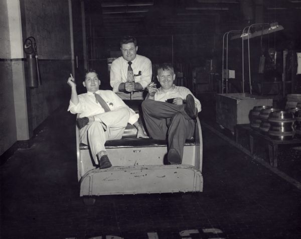 Sid and two colleagues sitting on a seat on the factory floor. Sid and another man are sitting on the seat bench, and the other men is sitting in the center behind them. The man on the left is holding a cigarette, and Sid is holding a film holder in his left hand and a cigarette in his right.