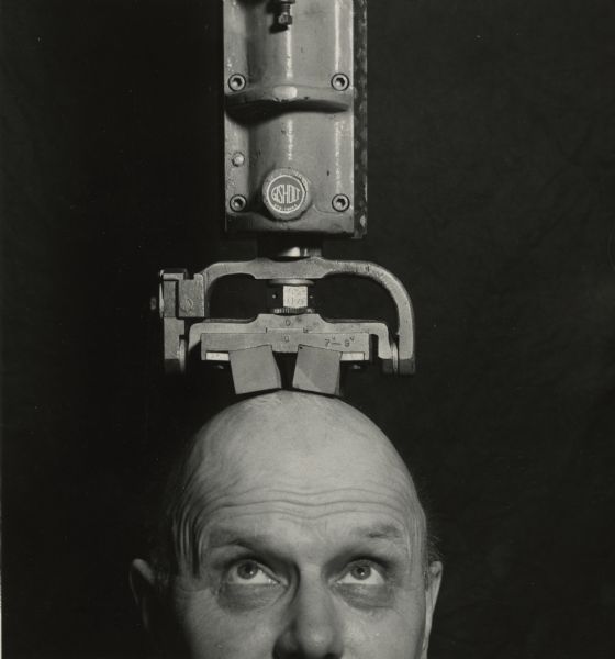 Funny portrait of a man looking up at a Gisholt machine resting on top of a his bald head. Below the round Gisholt trademark is the number 372-7306A.
