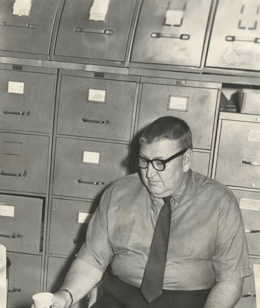 Gisholt office worker sitting in front of a bank of file cabinets, holding a paper cup in his hand. He is wearing a short-sleeved shirt with a button-down collar and a solid tie.