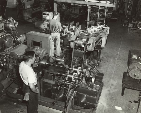 Elevated view of two Gisholt workers operating machinery on the factory floor. They are wearing t-shirts and jeans or khaki work pants; both men have rags in their back right pockets.