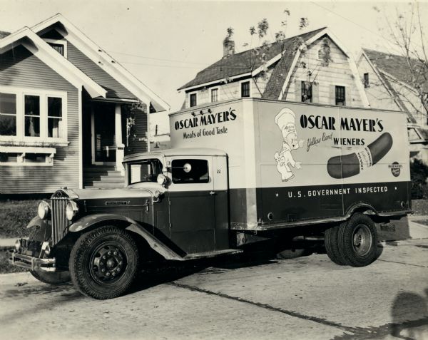 Delivery truck parked on the street of a residential neighborhood. The writing on the truck's front reads: "Oscar Mayer's/Meats of Good Taste." The writing on the side reads: "Oscar Mayer's Yellow Band Wieners/U.S. Government Inspected" along with an illustration of an cartoon character dressed as a food worker in white, and a yellow band wiener also appear on the left side.
