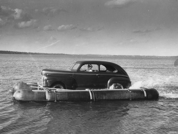 John "Commodore" Heggestad driving a car across Lake Monona on a pontoon. An article called "The 'Monona-Mobile'" was published in the Gisholt Machine Company employee newsletter <i>The News Crib</i> explains: "First he constructed a pontoon support for his car, then, on the rear tires, he chained wooden cleats which serve as paddles and propel the pontoon car at a respectable speed--as high as 10 knots per hour. Steering is accomplished by a rudder suspended from the rear axle. Gisholt lensman, Sid Boyum, who recently took an action photo of the skipper and his craft from the Lake Monona shoreline, reports that the 'cruiser' appears to be completely seaworthy."