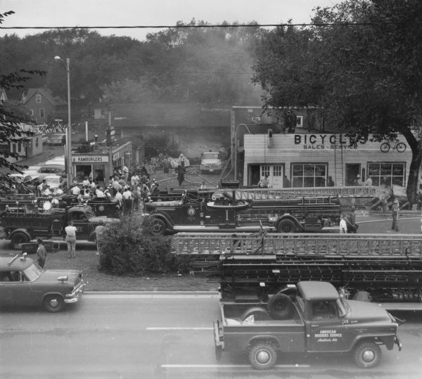 Elevated view from across street towards fire engines parked in front of Haack's Bicycles at 1250 East Washington Avenue. Firefighters are on the roof. There is a crowd gathered near a small restaurant with a sign for "Hamburgers."