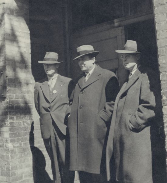 Three men standing in a doorway. All three men are wearing suits and hats, and two of them are wearing overcoats.