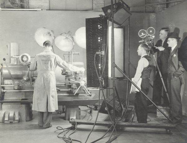 Three men are looking on, one running the movie camera, as a Gisholt worker is filmed working at a machine. The three men behind the camera are wearing suits or vests. The worker is wearing a long work coat with "Gisholt, Madison Wis." printed on the back. Large lights are behind the machinery.