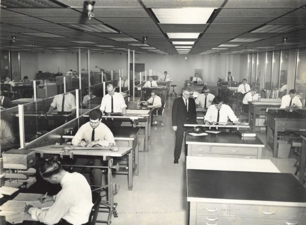 View down the center of a roomful of drafters working at their tables. Almost all the men are wearing white shirts and skinny black ties. A man in a dark suit is standing in the aisle near one of the draftsmen. A glass partition separates another room on the left, where people are also working at drafting tables. Probably  a drafting room at Gisholt Machine Company.
