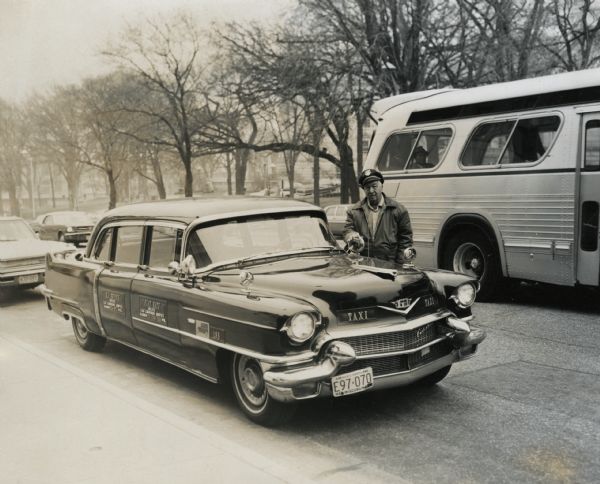 View from sidewalk of Bob Bender standing near the driver's side door of his dark 1950s model Cadillac taxi on the Capitol Square. He is wearing a leather jacket and a dark service cap. Signs on both right doors advertise his business, "R.L.  Bender Deluxe Cab Limousine Service" with a phone number. His cab number 198 is also visible. A bus is going by in the background. The trees on the Square in the background have bare branches. 