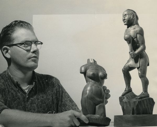 Robert Overman Hodgell, wearing a paisley shirt and eyeglasses, contemplating two wooden sculptures. The sculpture on the left is a headless, armless naked female torso; the one on the right is an apparently "tribal" figure.