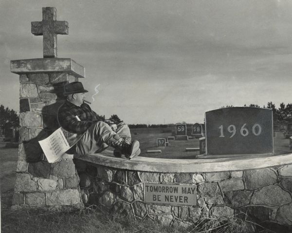 Sid is sitting on a stone wall looking out over a graveyard. He is wearing a hat and a checkered hunting jacket and has a cigar in his mouth. A stream of fake smoke is rising from end of the cigar. A Union Pacific Railroad calendar opened to July 1961 is under his right arm. "57," "59," and "58" are on headstones in the background; the headstone bearing the year "1960" is in the foreground. The stone cemetery wall bears the aphorism: "Tomorrow May Be Never."