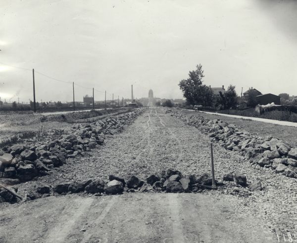 Construction work, probably on East Washington Avenue. The Wisconsin State Capitol is in the far distance. A farmhouse is on the right. A pickax is in the foreground near a pile of rocks. 