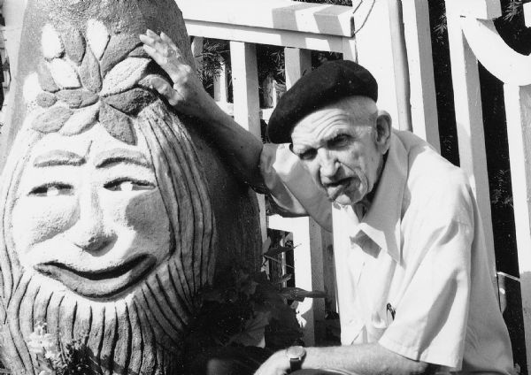 Sid posing with the "Gnome" sculpture in his backyard, hand resting on its head. Holding a signature cigar in his mouth, he is wearing a dark beret, a short-sleeved white button-down shirt, and a watch on his left wrist. A pair of reading glasses are in his breast pocket. The composition frames their two faces. 
