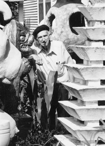 Sid standing in the backyard on Waubesa Street among his forest of concrete cast sculptures, like the "Multi-Tiered Pagoda," "Blue Tripod," and "Biomorphic Form." Holding a cigar in his mouth, he is wearing a dark beret, a short-sleeved white button-down shirt, dark trousers, and a ring on his right ring finger. A corn plant is directly in front of him.