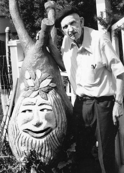 Sid standing with the "Gnome" sculpture in his backyard. Holding a cigar in his mouth, he wears a dark beret, a short-sleeved white button-down shirt, dark trousers and belt, and a watch on his left wrist. He has a pair of reading glasses in his left breast pocket.