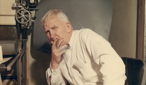 Sid posing next to the print dryer in the printing room near the darkroom at Gisholt Machine Company. He has his signature cigar, holding it to his mouth. He is wearing a long-sleeved white shirt and plaid bow tie. Sid's fingernails are stained, probably with pyrogallol, a staining agent used in the development of film.