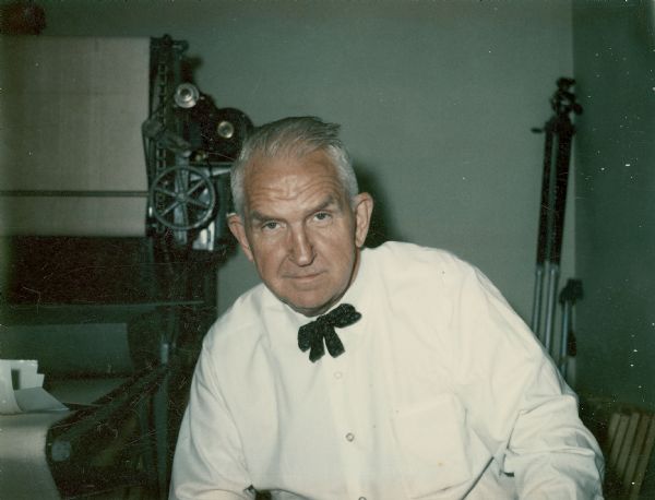 Sid posing in the printing room near a darkroom at Gisholt Machine Company. He is wearing a long-sleeved white shirt with a patterned bow tie. A print dryer is in the background.