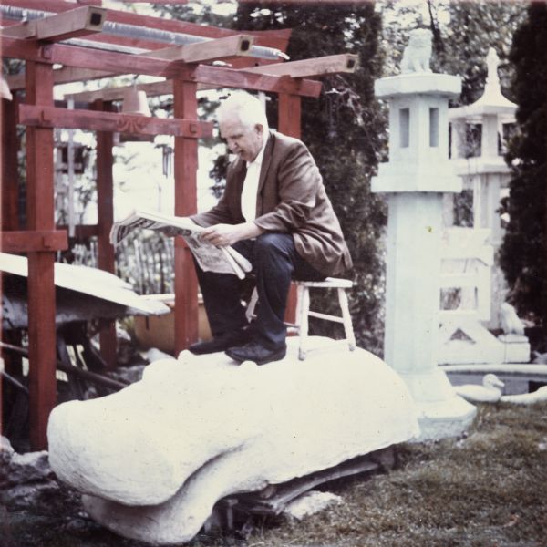 Sid sitting on a stool on top of his "Hippopotamus" sculpture in his backyard at 237 Waubesa Street. He has a cigar in his mouth and is reading a newspaper. Behind him are two lantern sculptures (one being the "Fu Dog Lantern"), shrubs, and a pond.