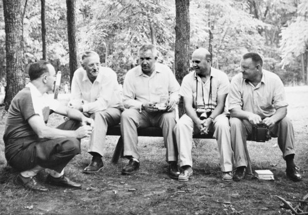 Sid and other four men sitting together on a bench under trees. Two of the men have cameras.